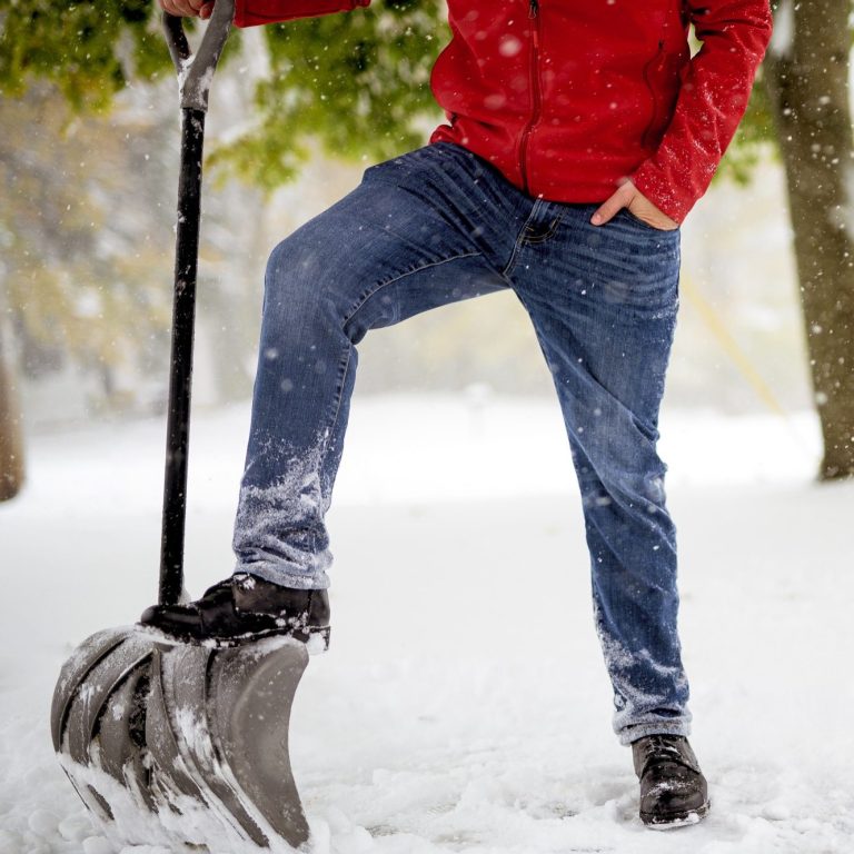 Person mit roter Jacke und Jeans steht mit einem Schneeschaufel im Schnee.